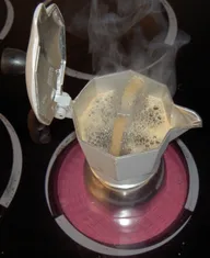 A Bialetti Moka Express brewing on an electric stove, lid open and steam rising