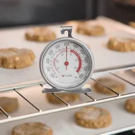 A Taylor oven thermometer standing on the rack of a lit oven among trays of cookies