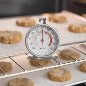 A Taylor oven thermometer standing on the rack of a lit oven among trays of cookies